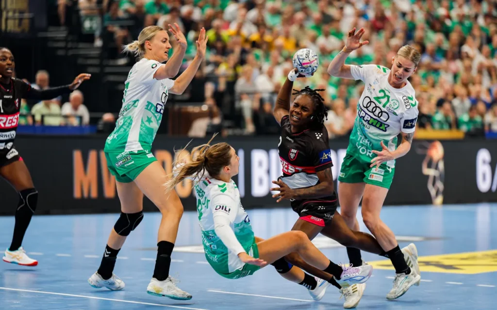 A handball player in a black jersey leaps to catch the ball while being defended by three opponents in green and white uniforms during an intense match.