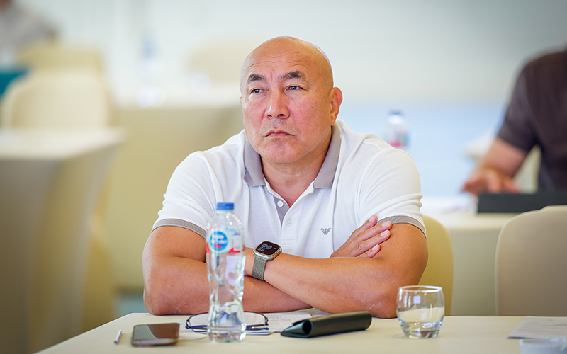 A person sits at a table with arms crossed, wearing a white polo shirt and a smartwatch, in a conference or meeting setting.